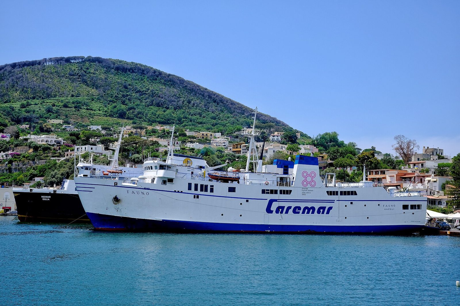 A white Caremar car ferry moored in a harbor on Ischia with green hills behind