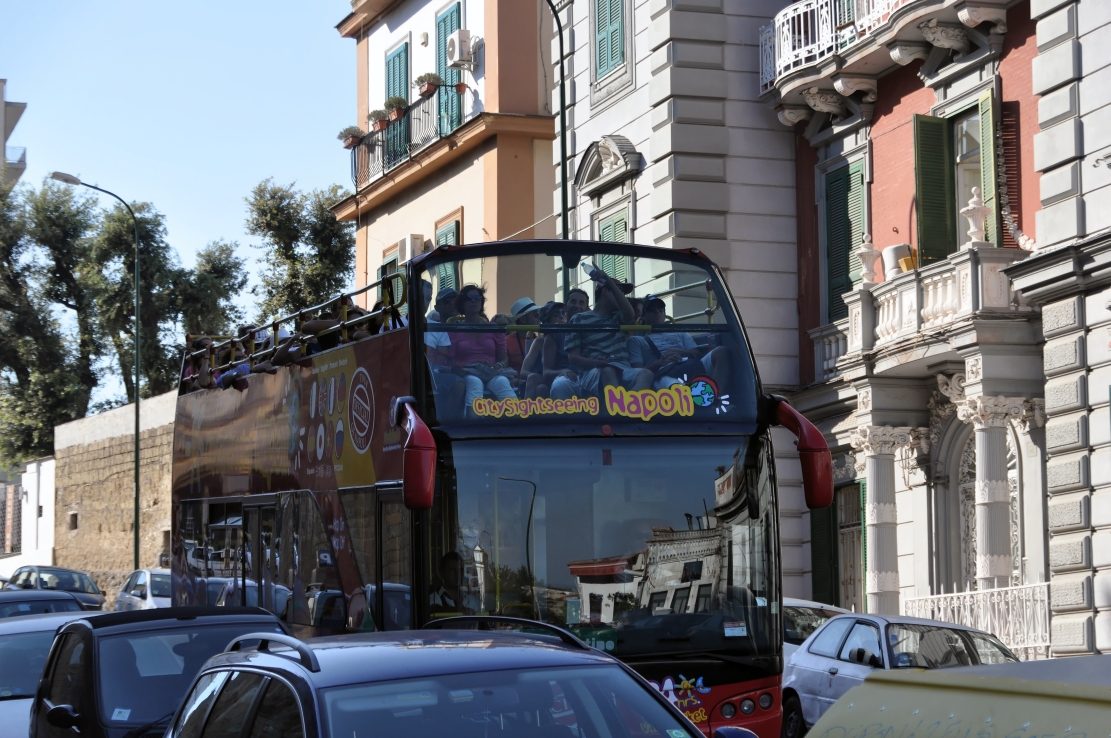 An open-top hop-on hop-off sightseeing bus in traffic on a Naples street