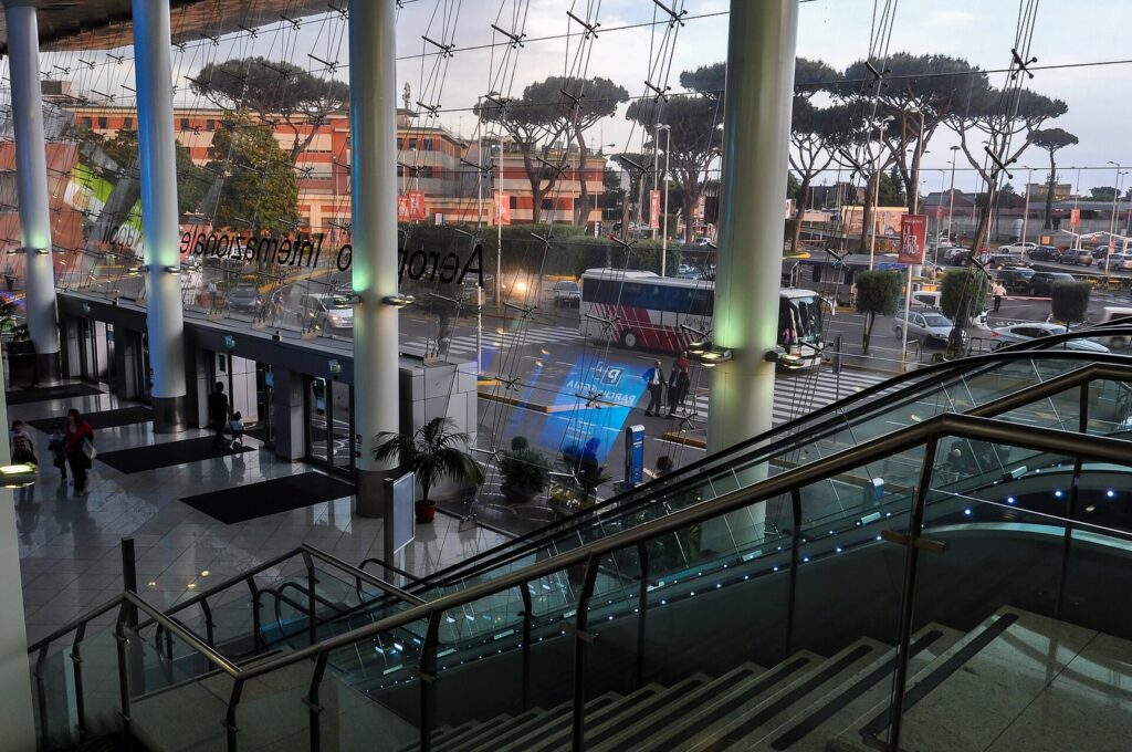 Inside the Naples Capodichino airport terminal, with escalators and umbrella pines visible through the glass