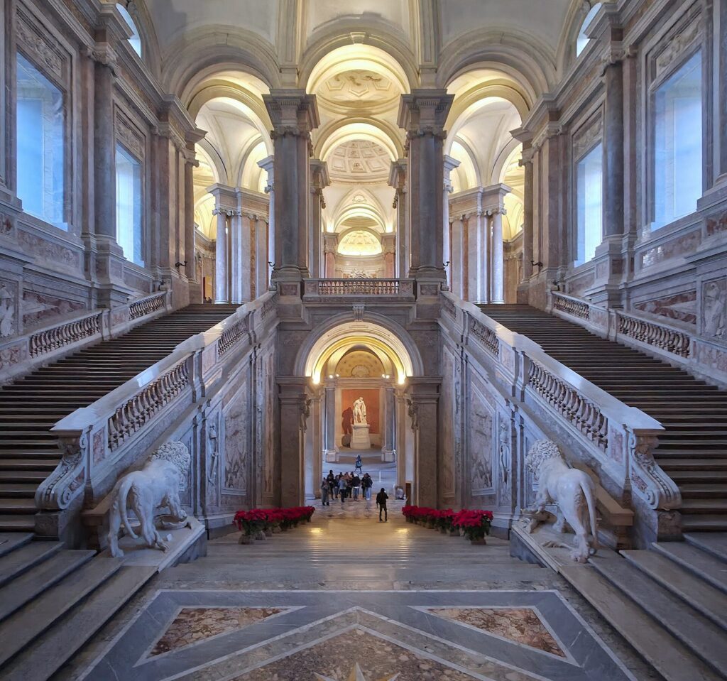The marble grand staircase of the Reggia di Caserta rising between stone lions under a painted vault