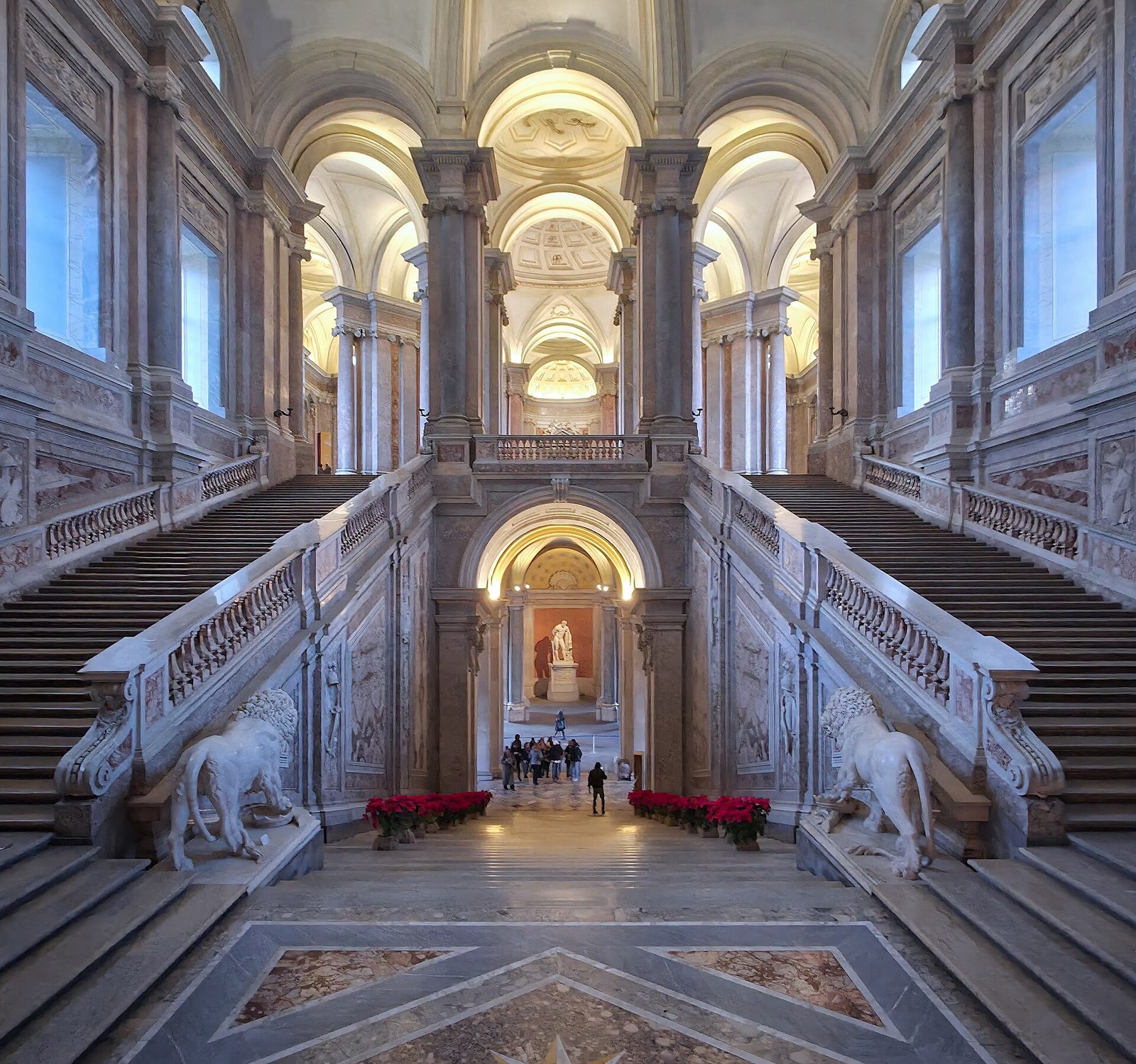 The marble grand staircase of the Reggia di Caserta rising between stone lions under a painted vault