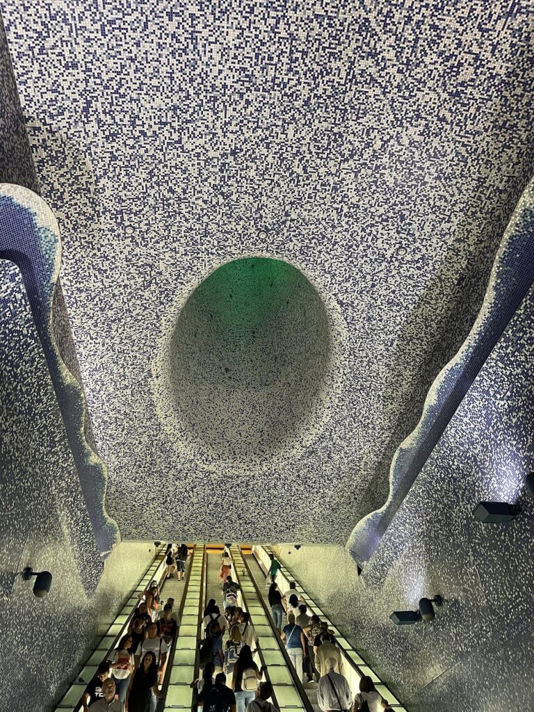 Escalators rising toward the blue mosaic crater of light at Toledo metro station in Naples