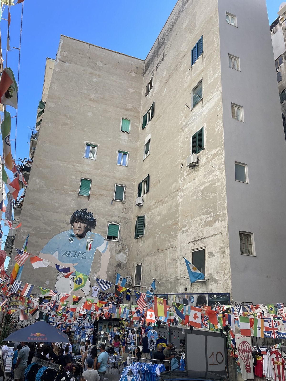 Large mural of Diego Maradona on a tall building in the Quartieri Spagnoli, with flags and visitors below