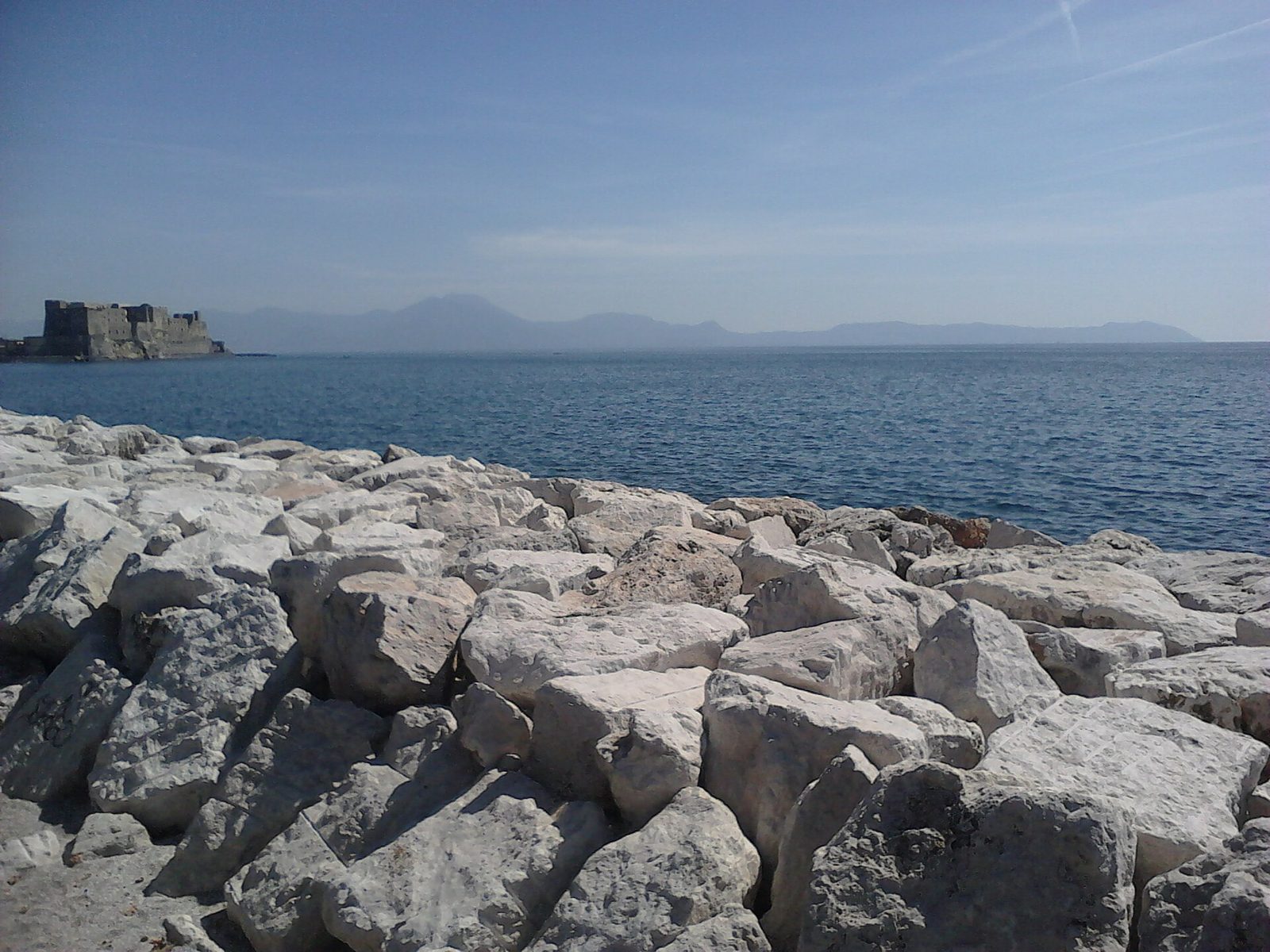Rocks along the Naples seafront with Castel dell'Ovo and the bay behind under a clear sky